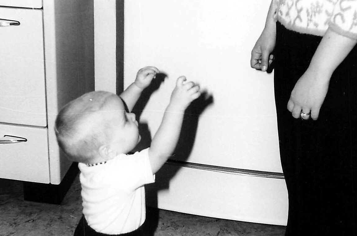 Young child reaching up toward an adult's hands near a refrigerator in a kitchen, daytime scene (black-and-white photo). When in doubt, concise and descriptive of action and setting.