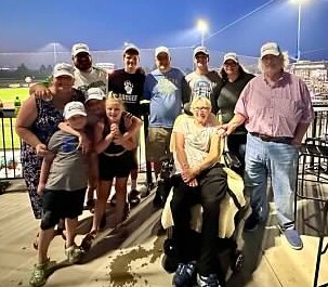 Group of people posing together on stadium bleachers at night, with a person in a wheelchair at the front center.