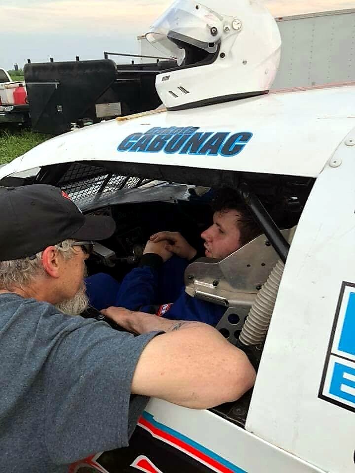 Racing driver sits in the open cockpit of a white race car while a man outside helps him buckle up. Helmet resting on the car’s roll cage above the cockpit.