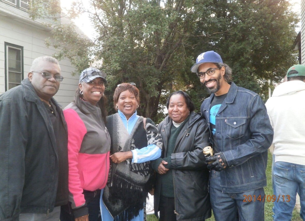 Group of five adults posing together outside, smiling, in casual jackets, with trees and a house in the background. May include a visible date stamp of 2014/09/13 in the corner.