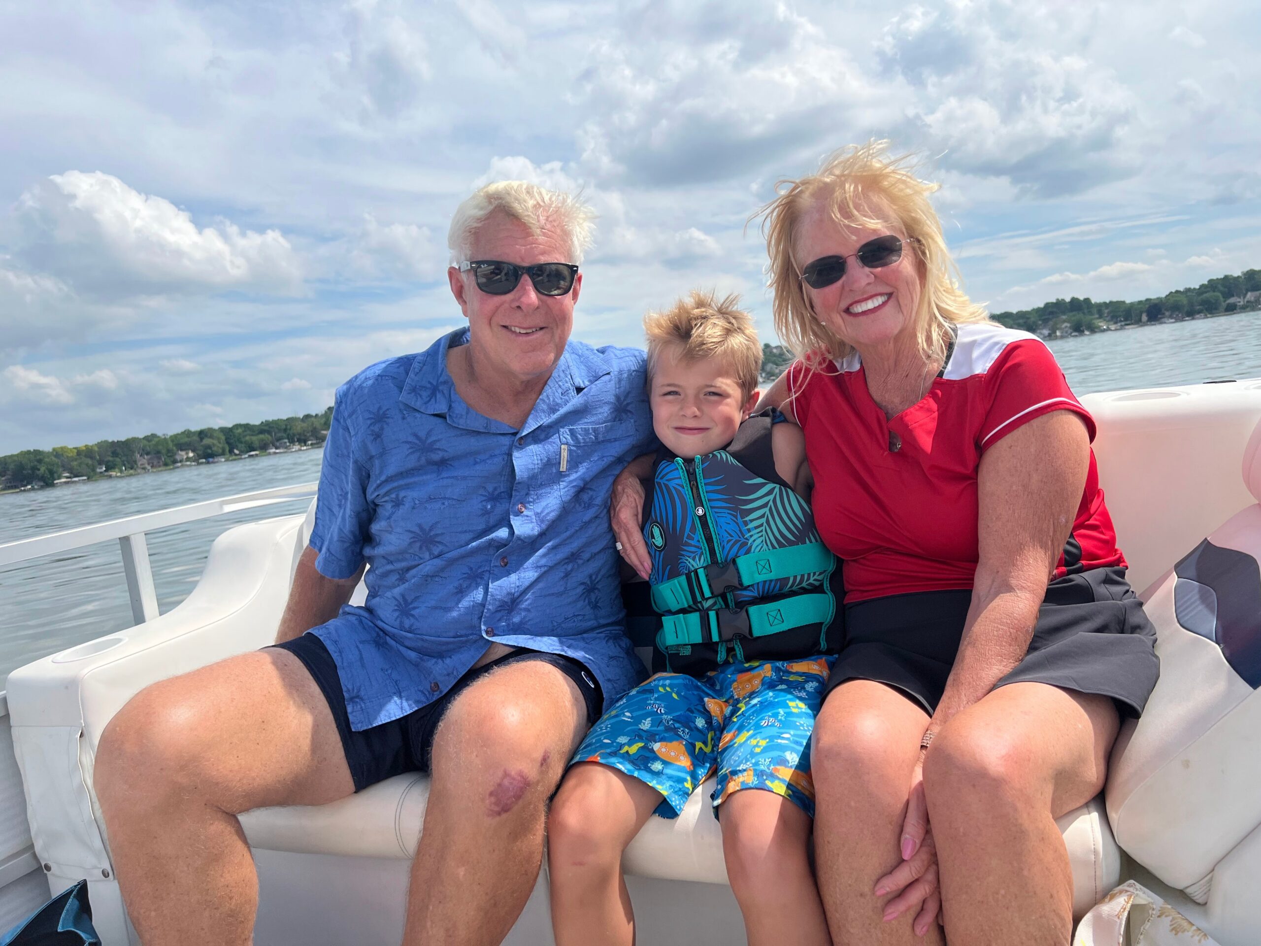 Family of three smiling on a boat, wearing life jackets, with water and shoreline in the background.