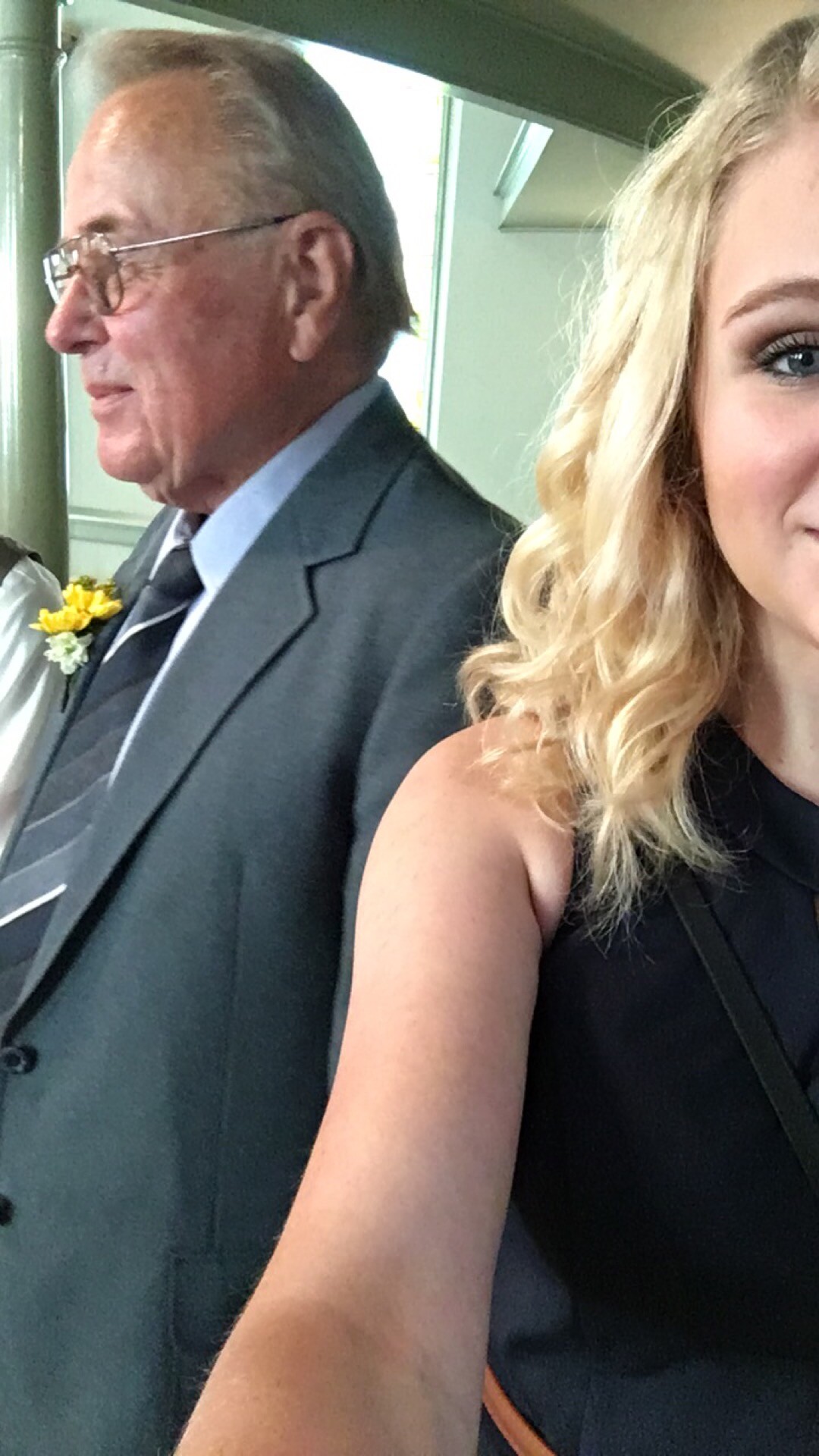 Older man in a gray pinstripe suit with glasses and a yellow boutonniere, standing beside a smiling blonde woman in a black dress at a formal event.