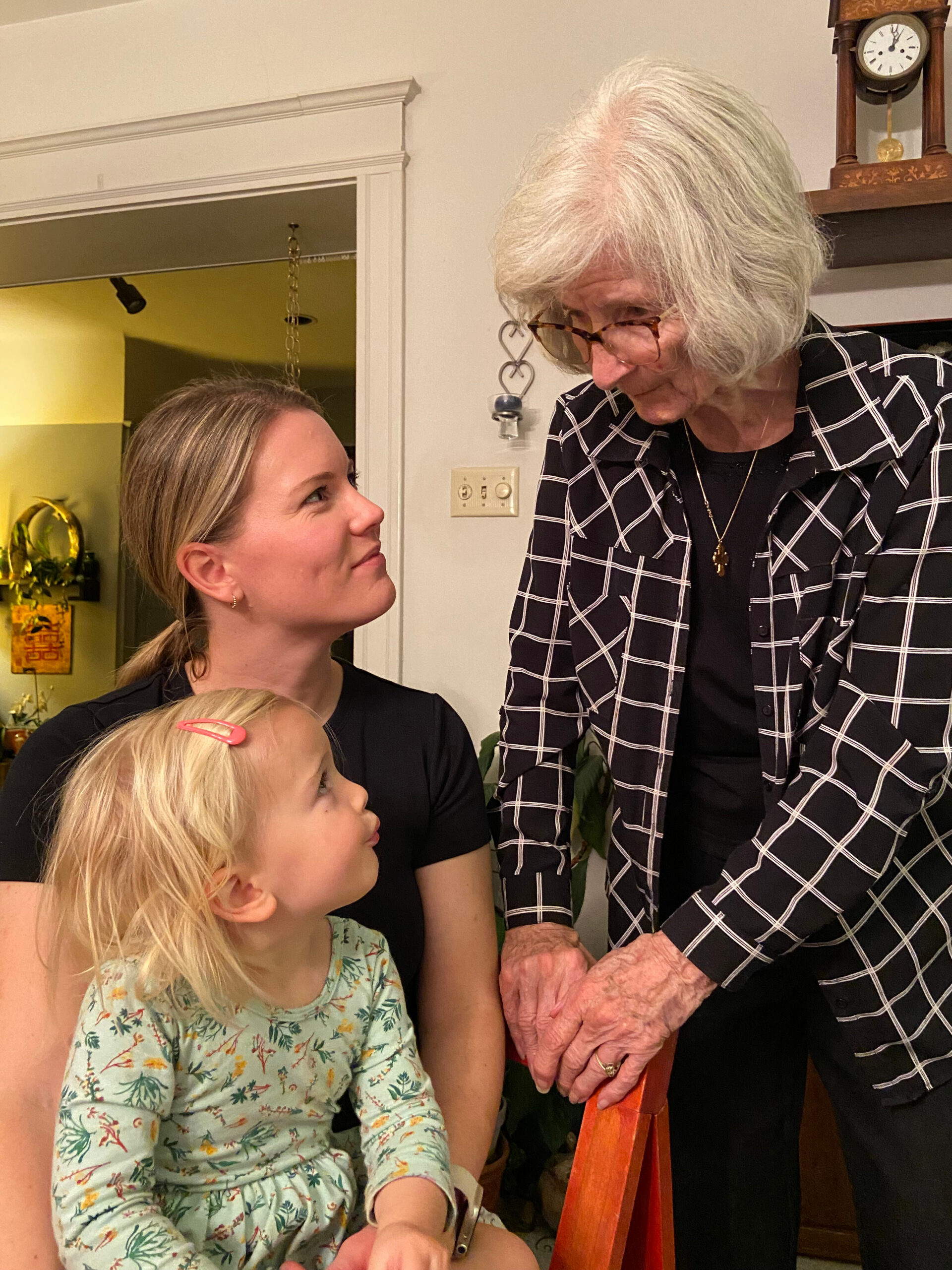 Elderly woman with white hair and glasses leaning toward a seated woman and child at home, engaging in a conversation in a living room.