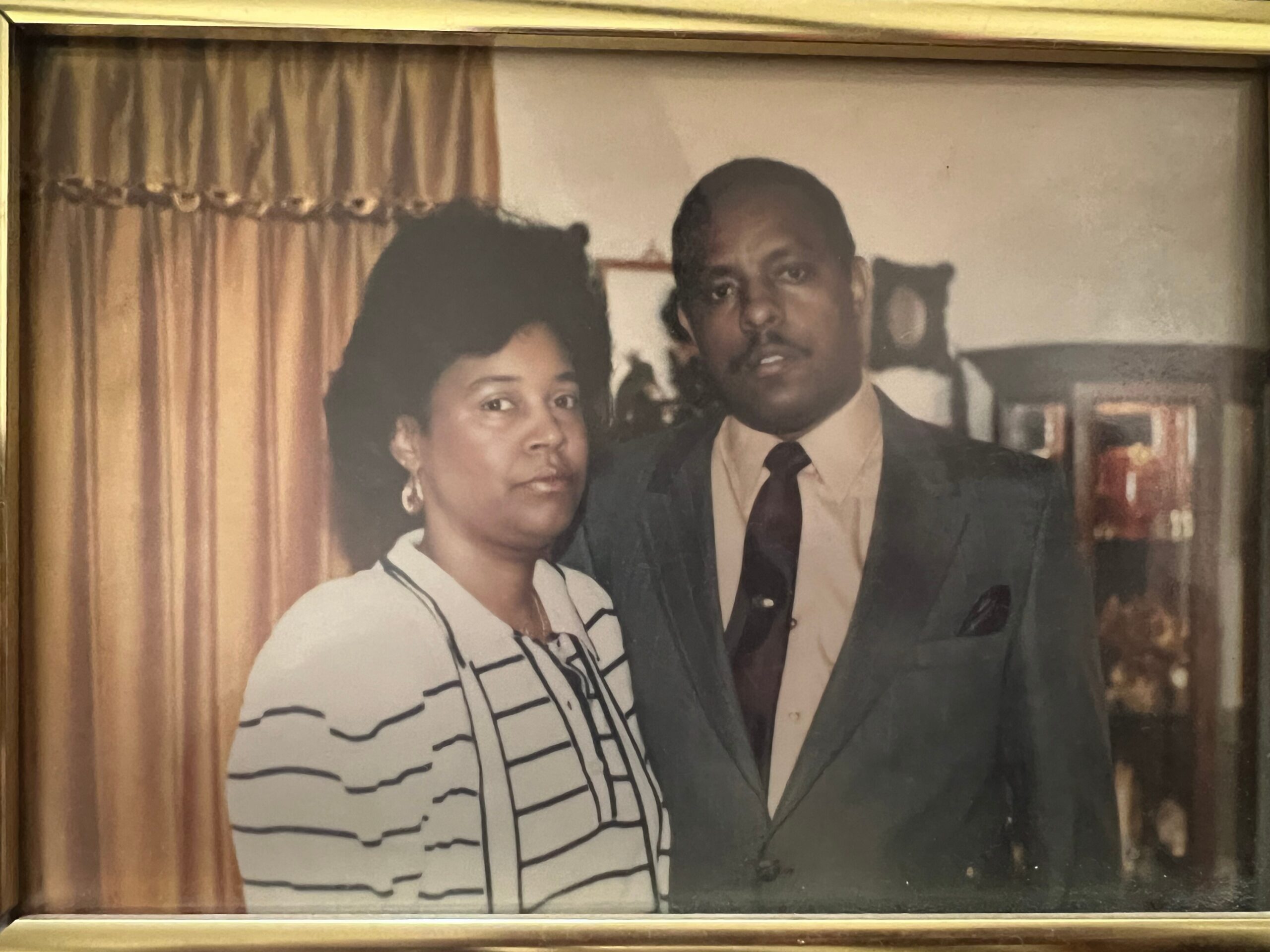 Portrait of a Black couple indoors; man in a suit and tie, woman in a striped top, standing together.