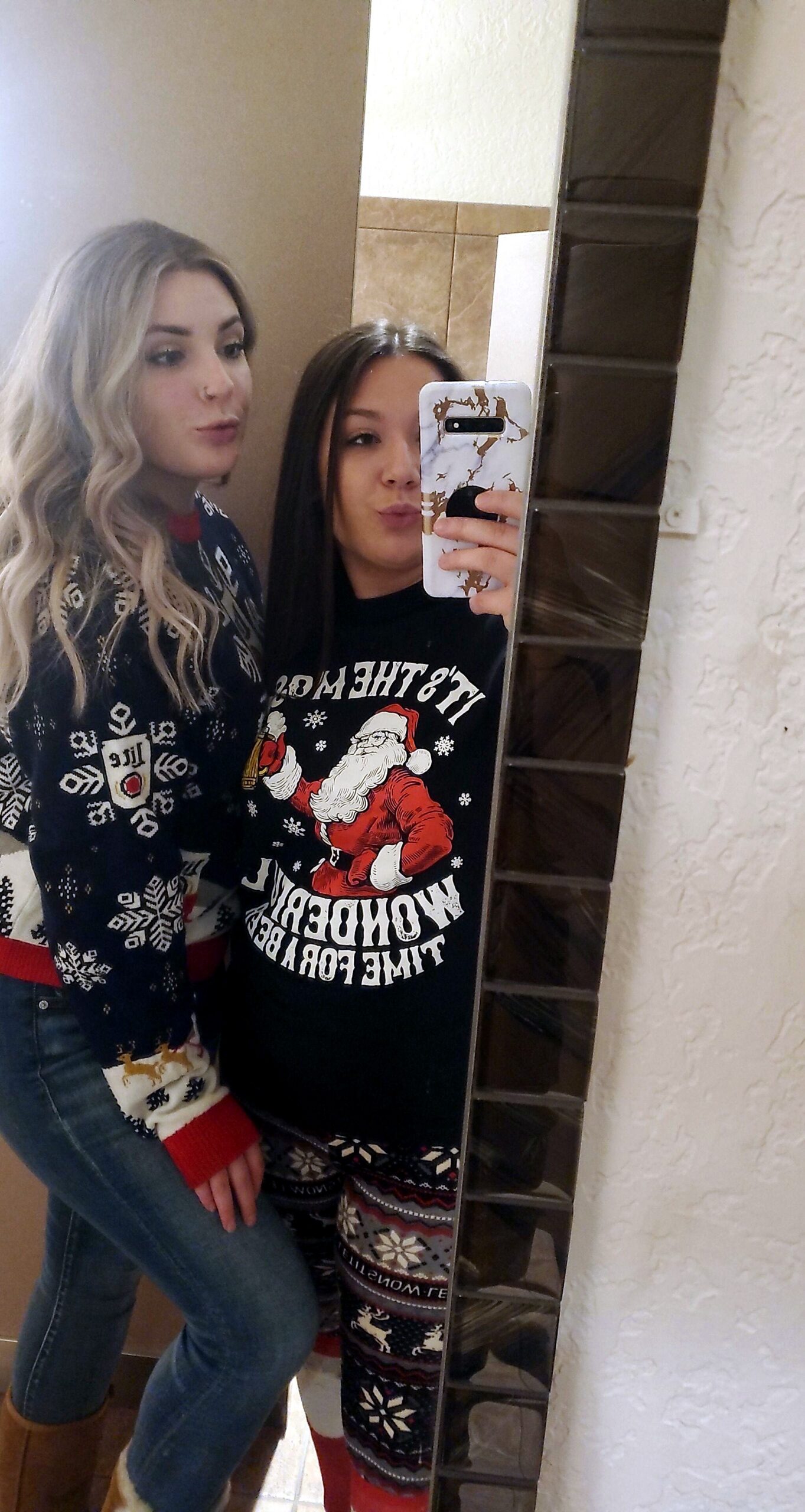 Two young women take a mirror selfie in a bathroom; one with blond waves wears a blue Christmas sweater, the other with dark hair wears a Santa sweater and patterned leggings, holding a phone.