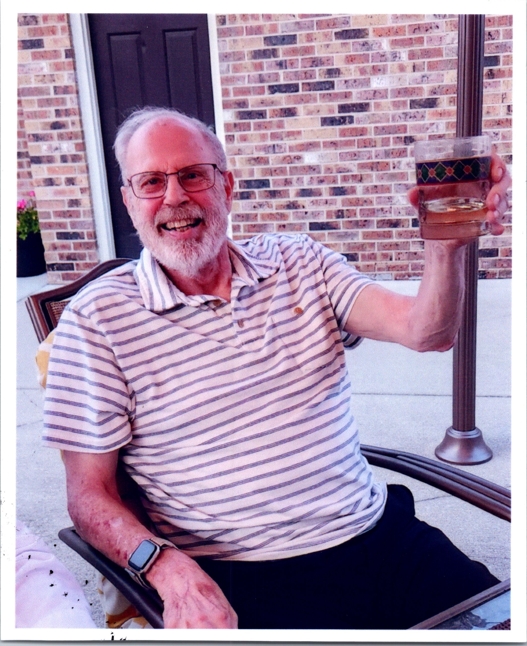 Smiling elderly man with glasses and white beard, in a striped polo, sitting on a patio and holding a decorative glass with a drink.