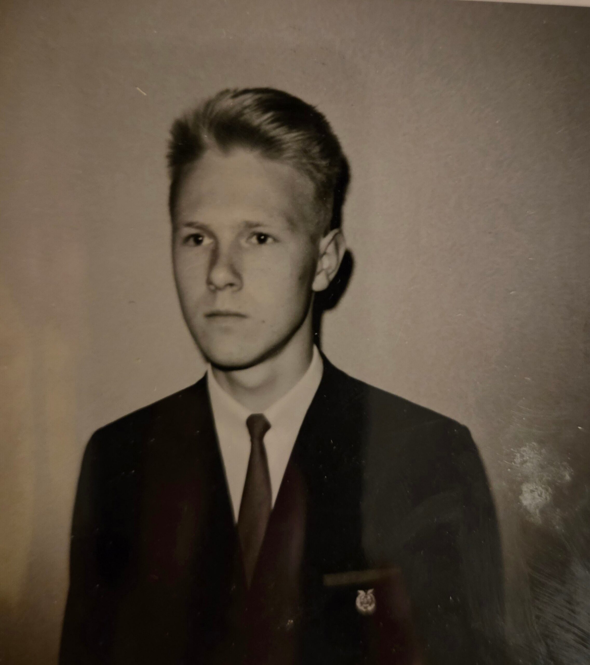 Young man in a dark suit and tie, formal studio portrait with neatly styled hair and a lapel pin on the jacket edge.
