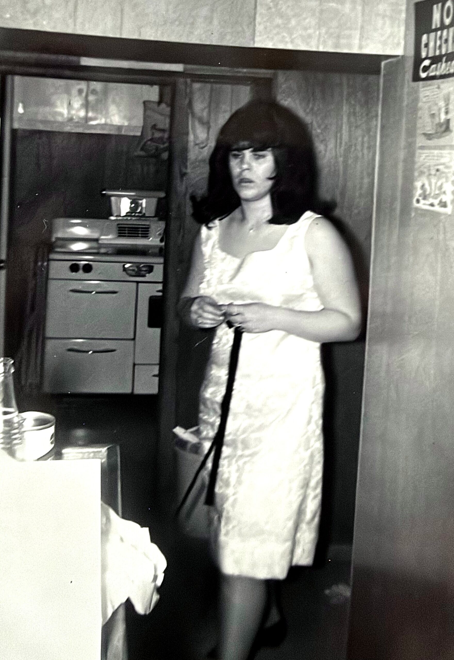 Black-and-white photo of a woman with dark shoulder-length hair wearing a light sleeveless dress, standing in a kitchen near a stove and doorway, looking toward the camera.