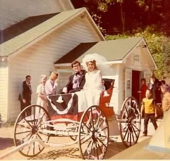 Bride in a white wedding dress sits in a red-and-white horse-drawn carriage in front of a small church, surrounded by guests on a sunny day.