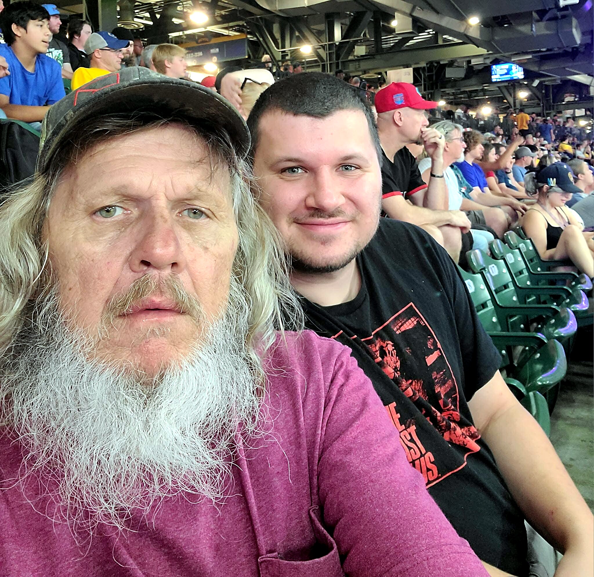 Close-up of two men taking a selfie in a crowded stadium, the bearded man in a purple shirt in the foreground and the smiling younger man beside him.