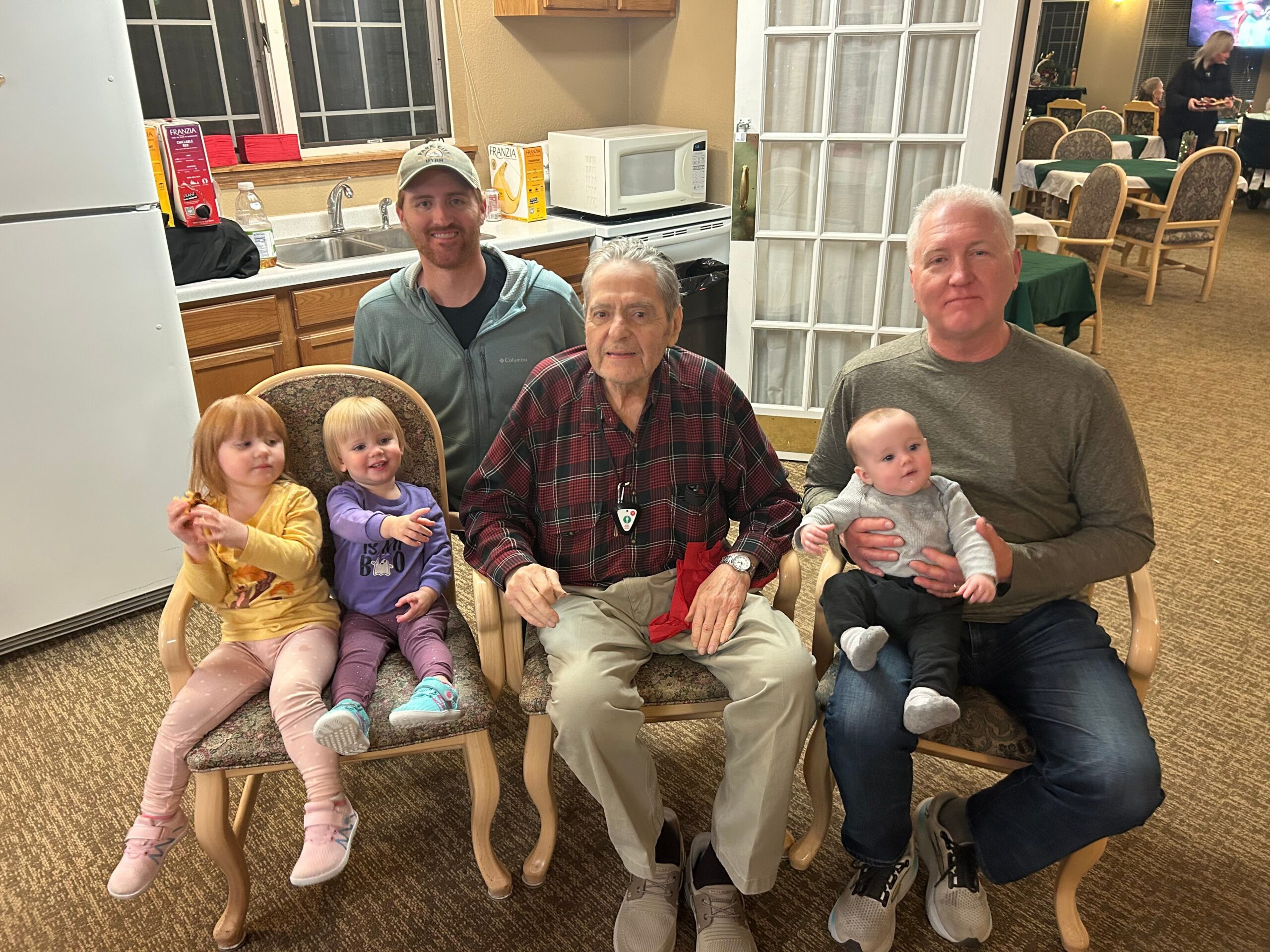 Group photo of three adults and three small children seated in a kitchen-like room at a social gathering.