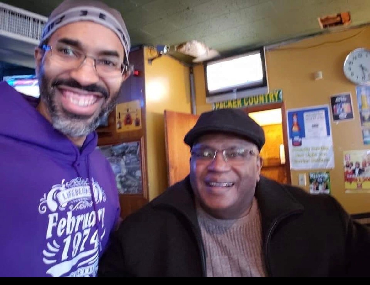 Two men smiling for a selfie indoors, one in a purple hoodie and headband, the other in a black cap and glasses in a cozy cafe setting.