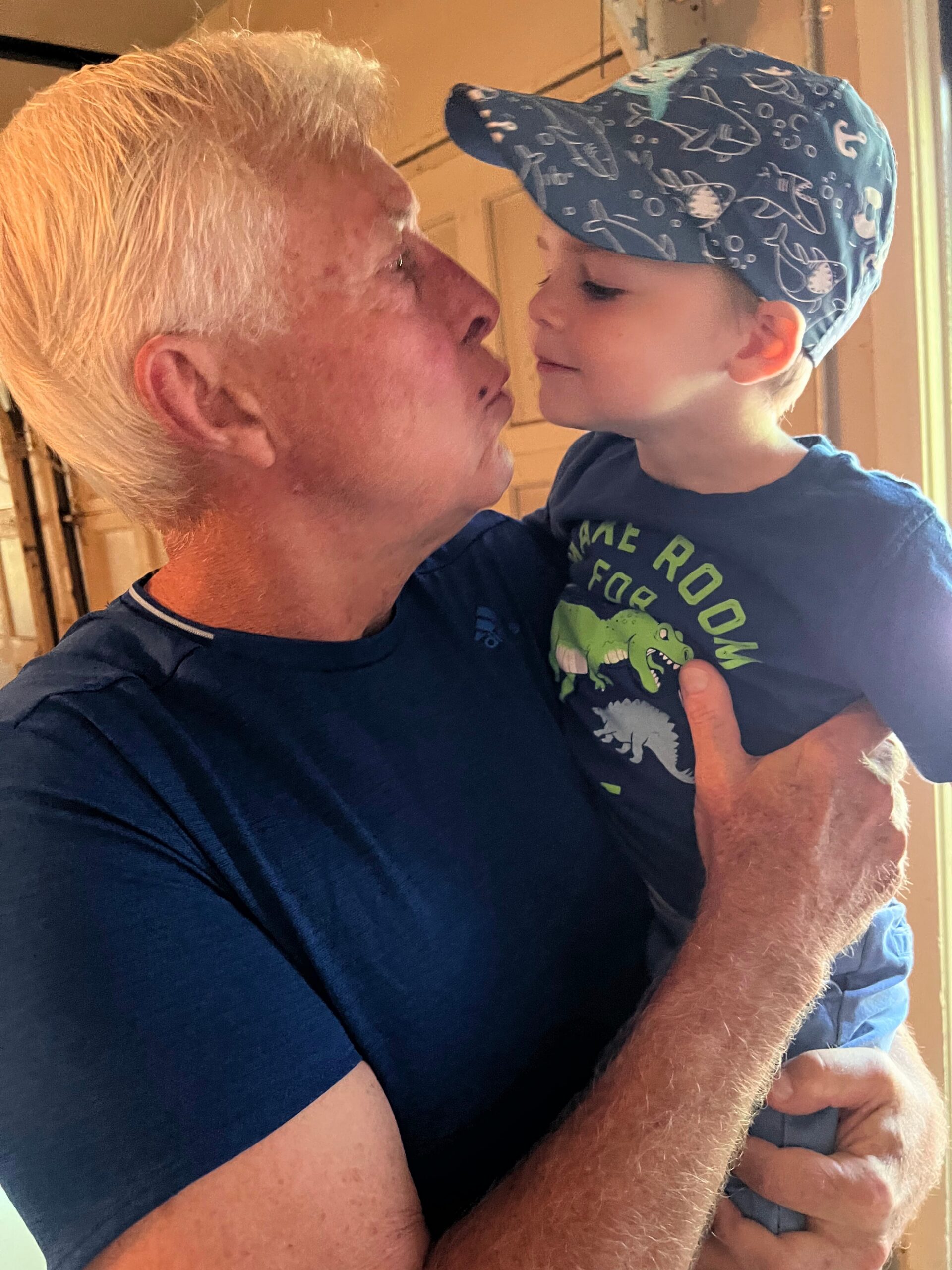 An older man holds a young child close as they touch noses in a loving moment indoors; the child wears a shark-pattern blue cap.