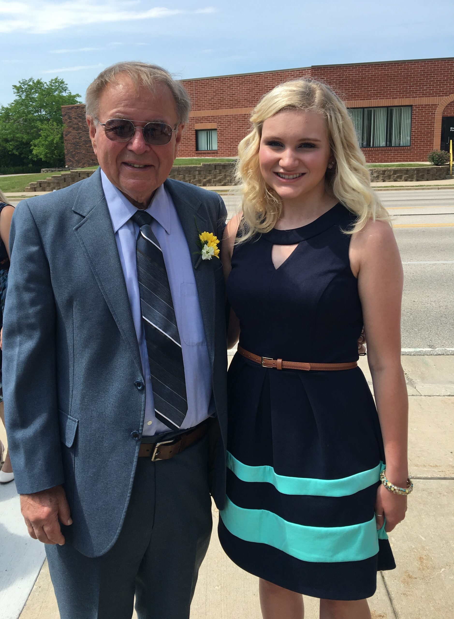 Older man in a blue suit and sunglasses posing with a young woman in a navy dress outside a brick building on a sunny day.