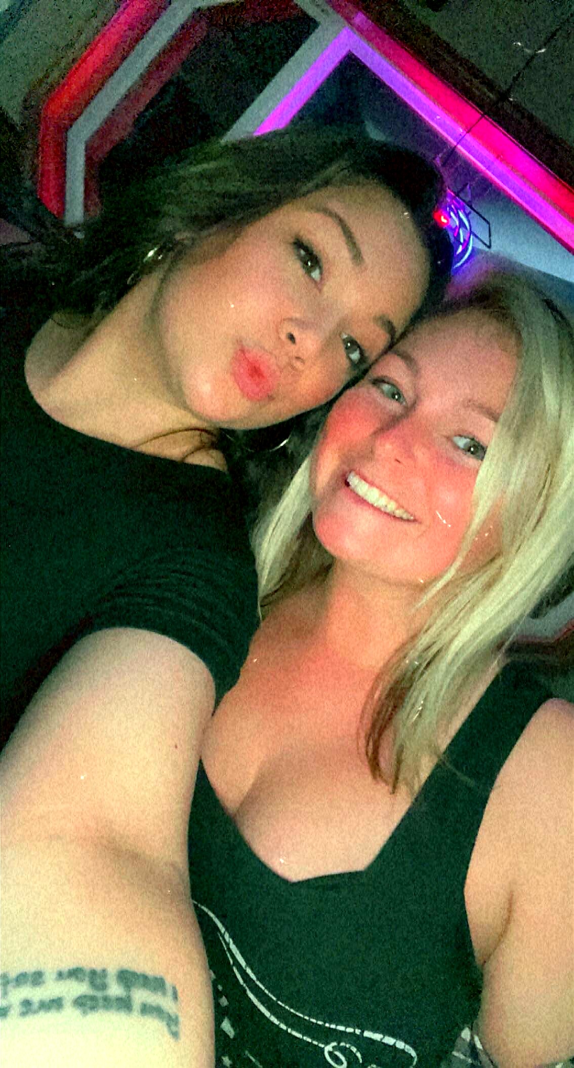 Two women posing for a selfie at a party; one puckers her lips, the other smiles, with neon lights in the background.