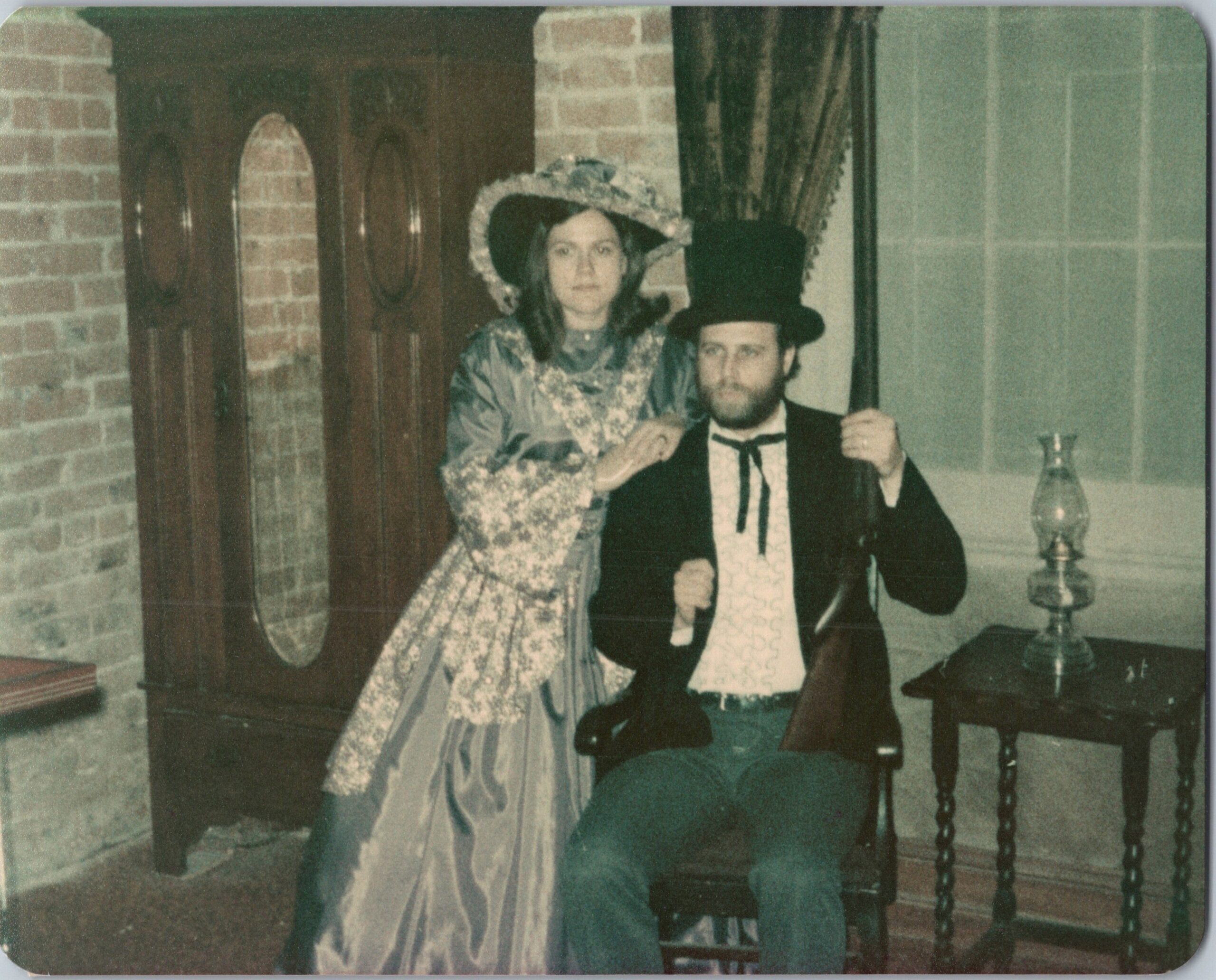Woman in a floral era dress and bonnet stands beside a man in a tuxedo and top hat at a brick-walled indoor setting, holding his arm.