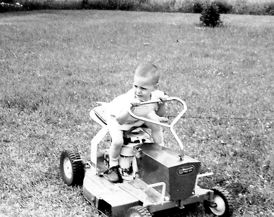 Baby riding a small ride-on lawn mower on a grassy field, gripping the steering wheel.