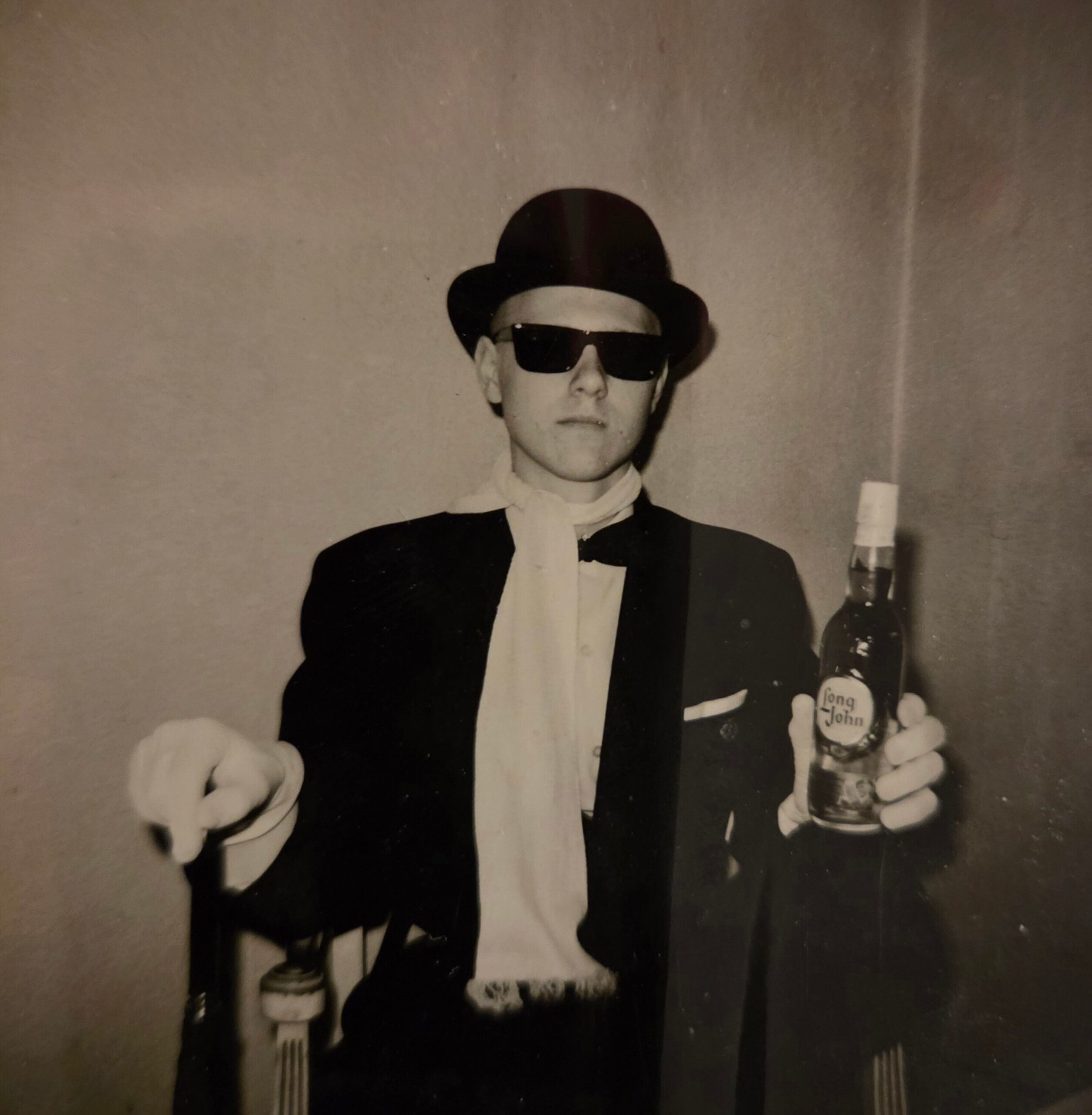 Black-and-white photo of a man in a tuxedo, top hat and sunglasses, holding a bottle and a fork.