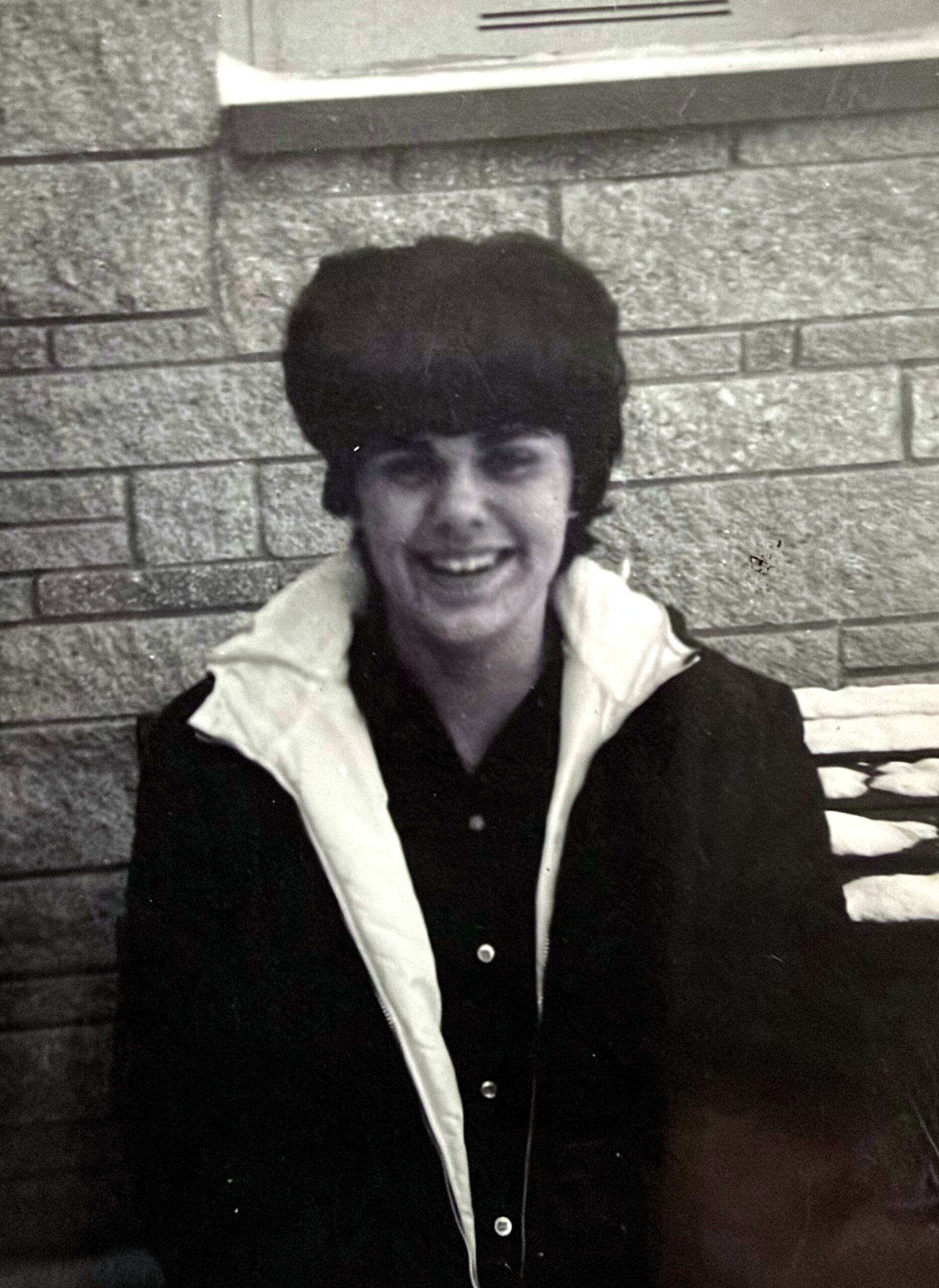 Smiling person with a large dark hairdo in a dark coat with a light collar, standing against a brick wall (black-and-white photo). 2-person portrait style placeholder.