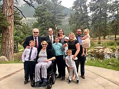 Group portrait of a multi-generation family outdoors in a park with mountains; includes a person in a wheelchair and several kids, all smiling.