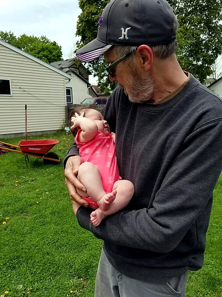 Older man in a dark cap and sunglasses cradling a baby in a pink onesie in a suburban backyard.