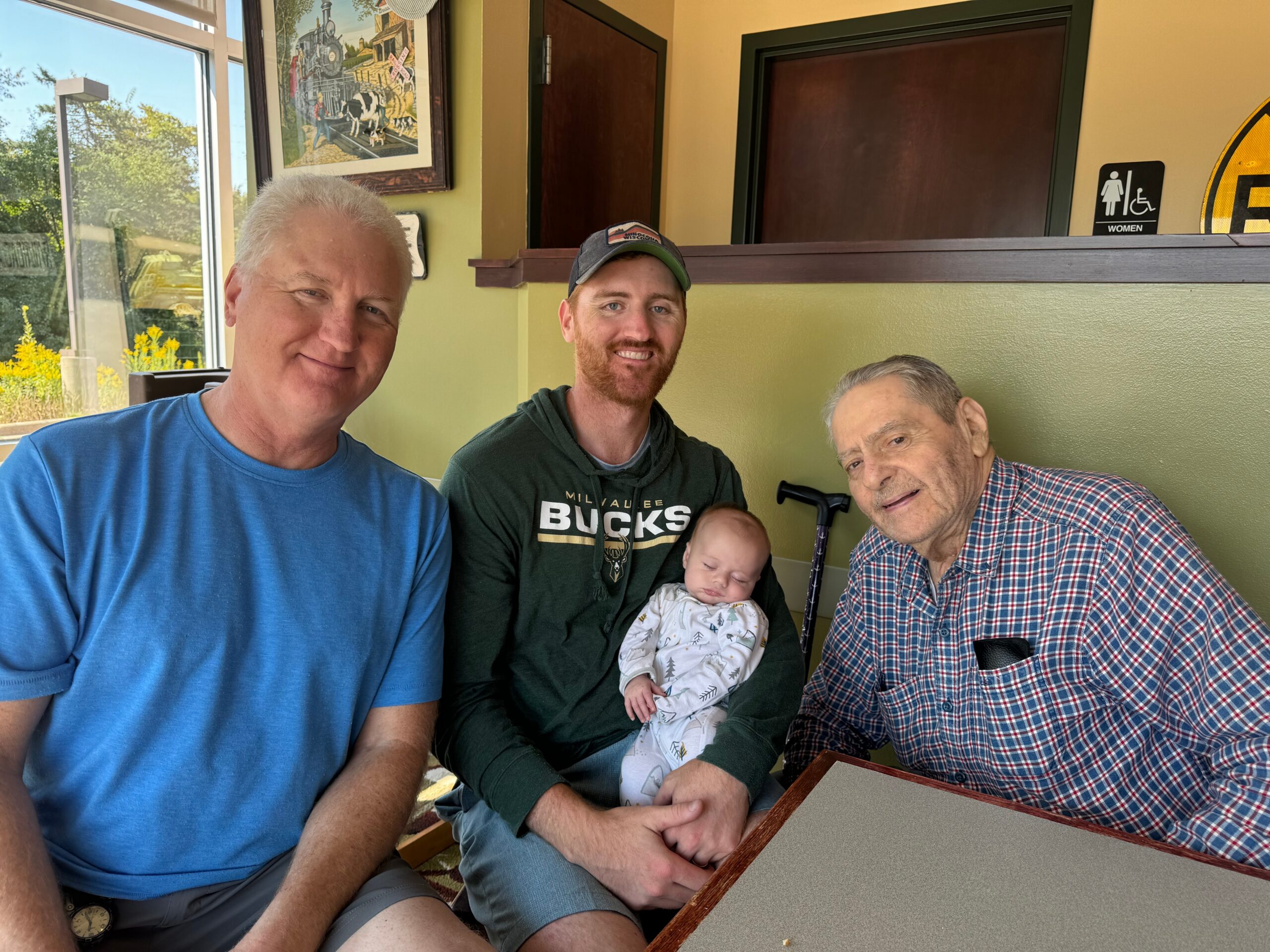 Group of three men and a sleeping baby posing for a selfie in a casual restaurant booth, smiling at the camera.