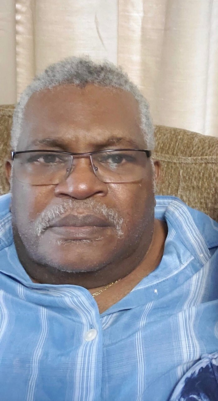 Close-up portrait of an older man with gray hair and glasses, wearing a blue shirt, seated on a sofa.