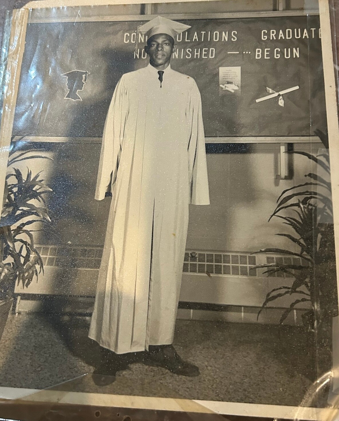 Black-and-white photo of a graduate in a cap and gown standing in front of a celebration banner that reads 'Congratulations Graduate'.