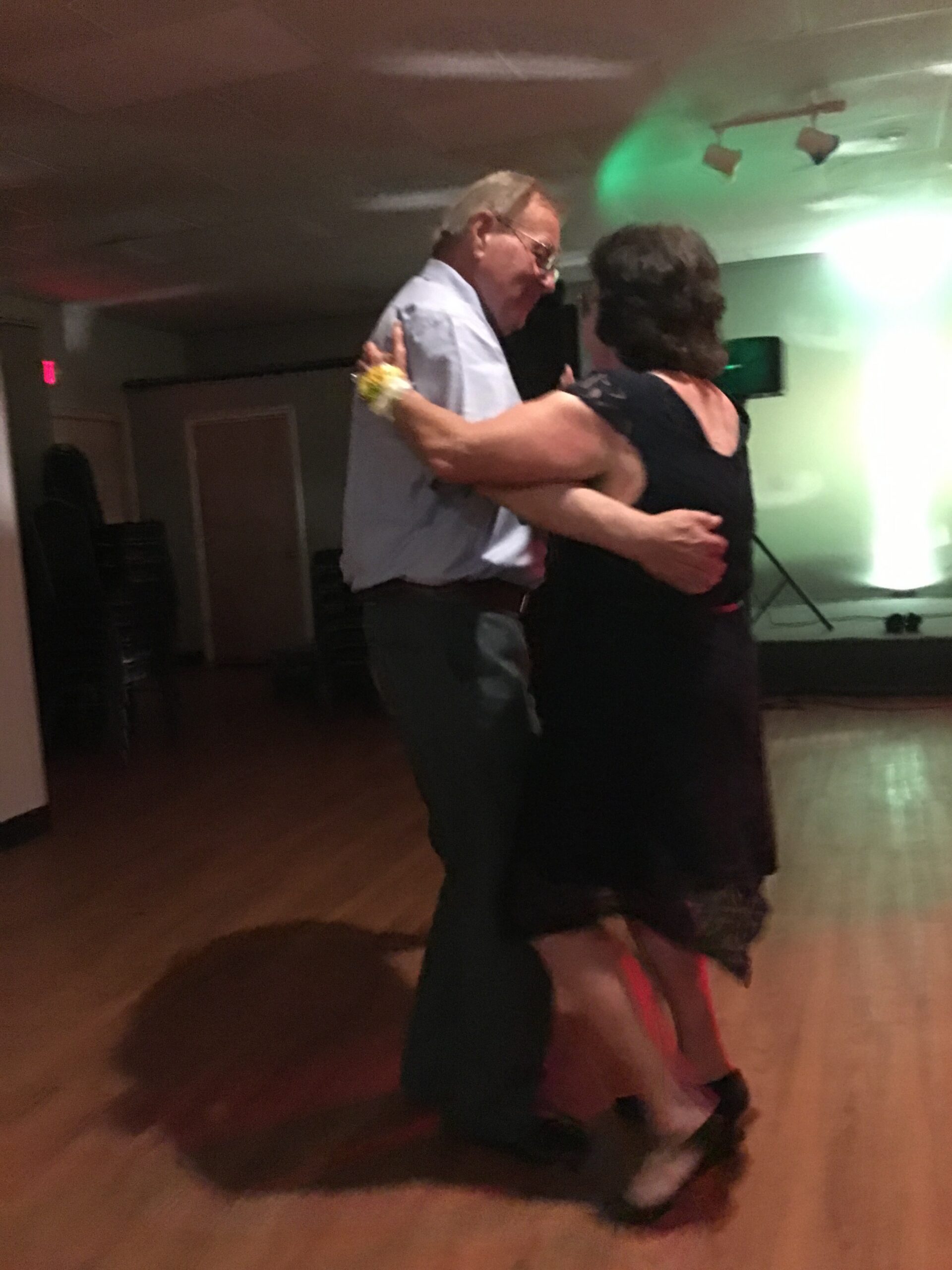 An older man in a light shirt dancing closely with a woman in a black dress on a dimly lit dance floor, both smiling.