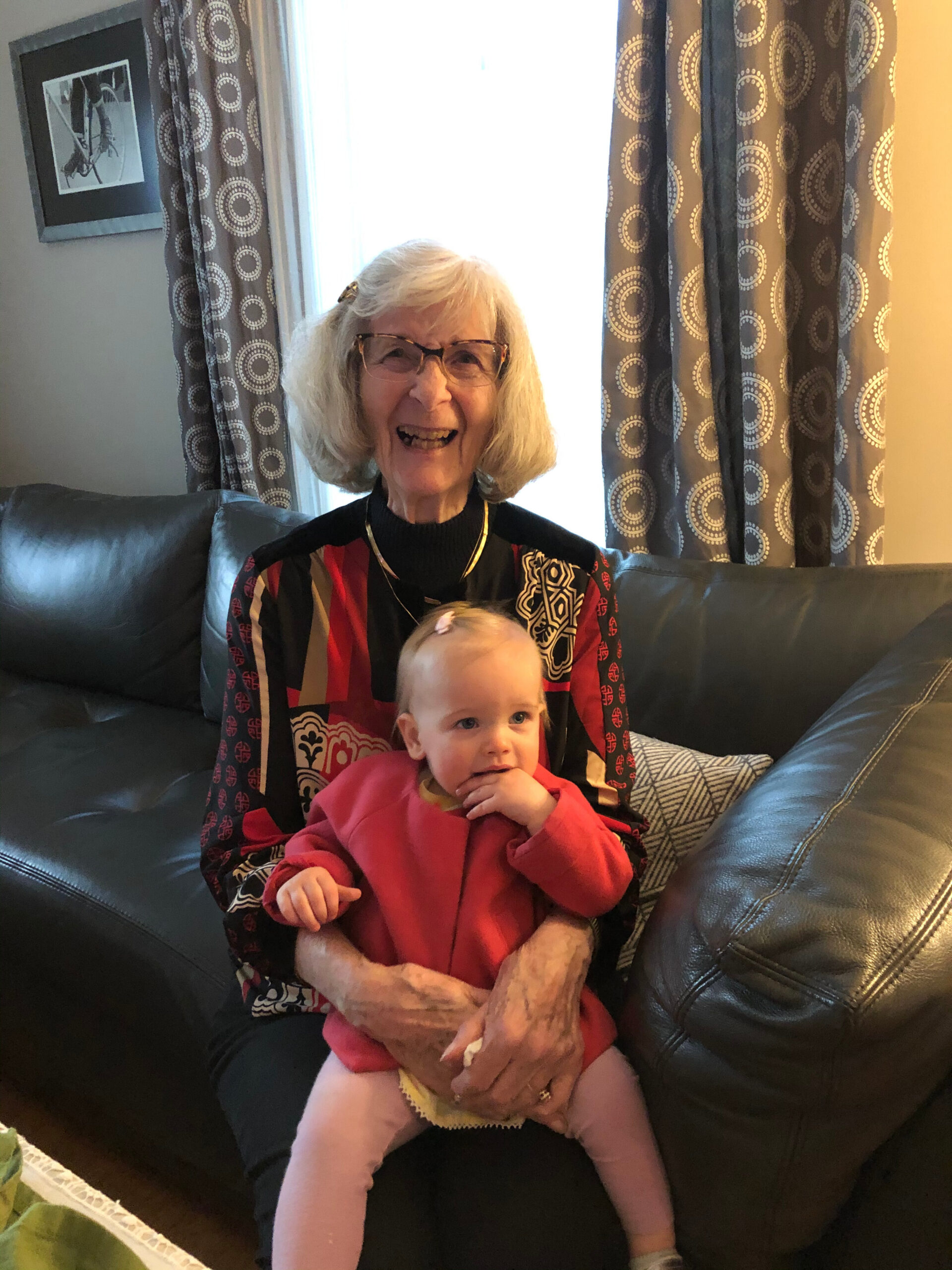 Smiling elderly woman with glasses on a black leather sofa, holding a baby in a red outfit indoors.