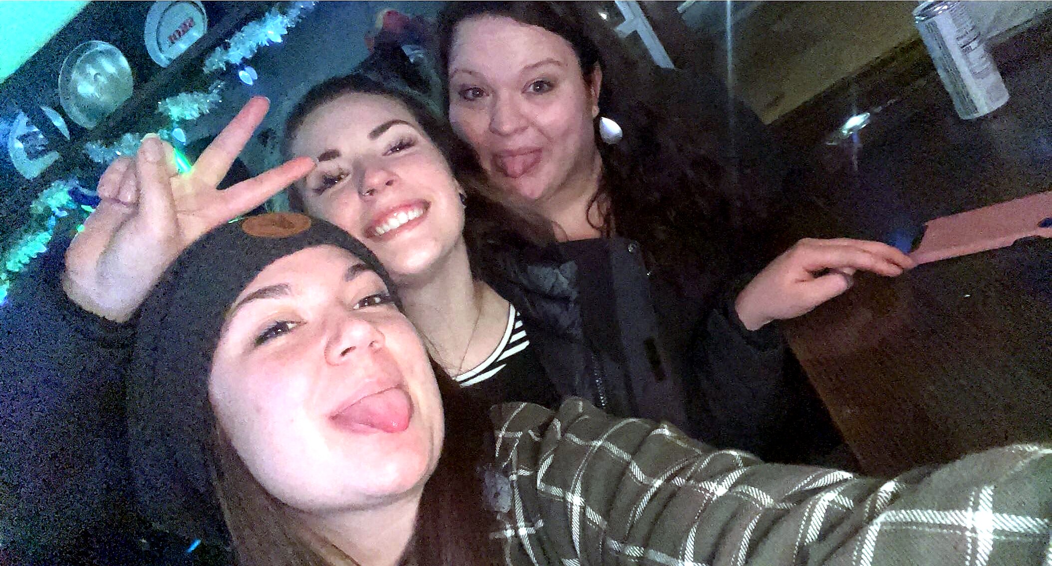 Three friends posing for a selfie in a dimly lit bar, smiling and sticking out their tongues.