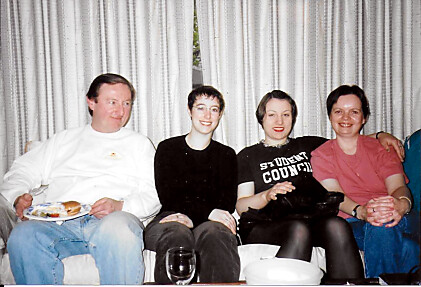 Four adults sitting on a couch, smiling at the camera, with drinks and snacks on the coffee table in front of them.