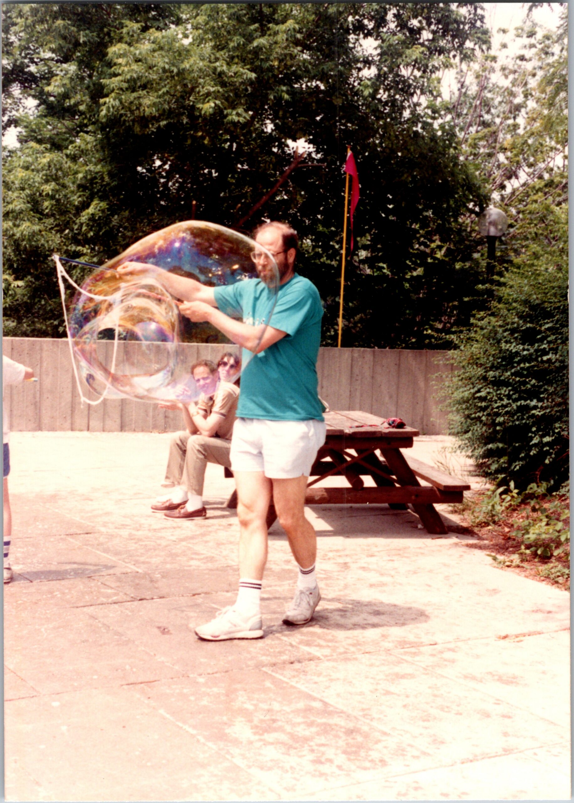 Man in a turquoise shirt and white shorts blows a giant soap bubble outdoors, while people sit at a picnic bench in the background.