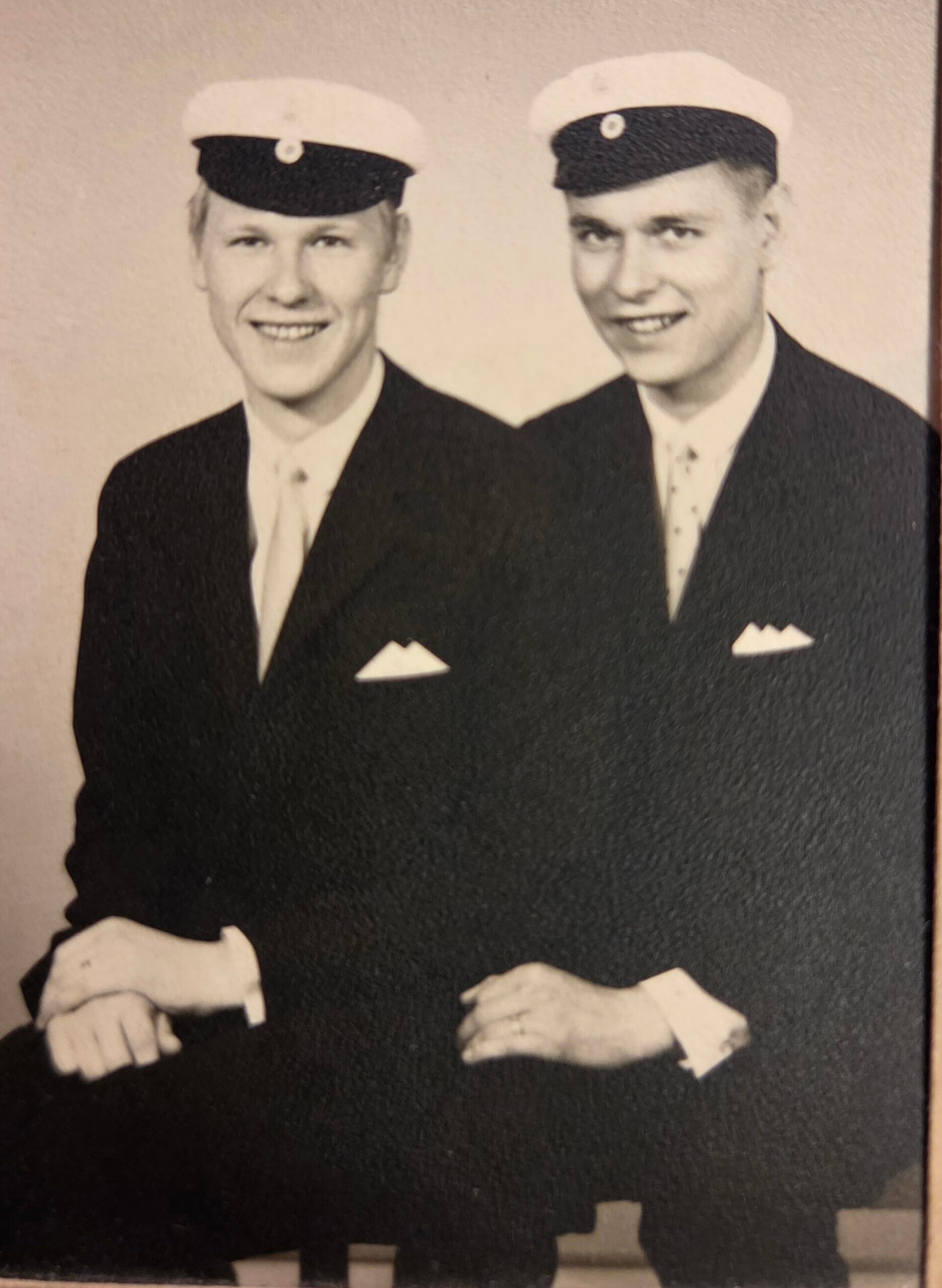 Two young men in dark suits and white sailor caps pose and smile for a photo.