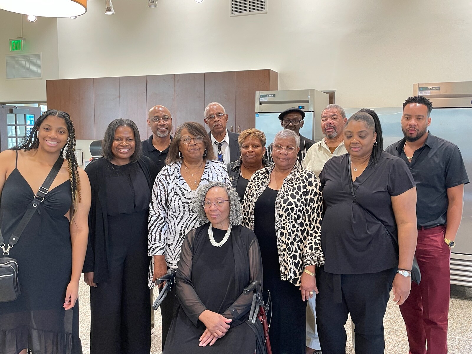 Group of twelve adults posing indoors for a group photo, most wearing black, with a woman in a wheelchair at the front center.