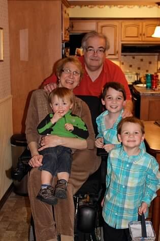 Smiling family in a kitchen: grandmother in a wheelchair holding a toddler, an older woman beside her, and two boys in front; a man in a red shirt stands behind them.