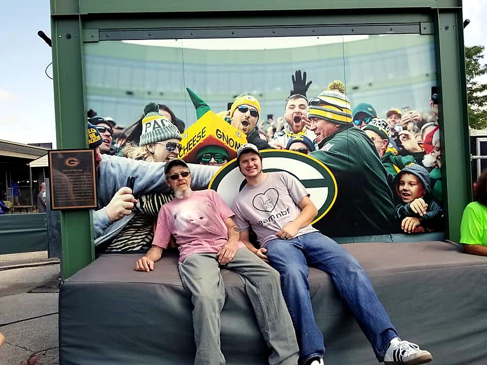 Two men pose on a bench in front of a large mural of cheering Green Bay Packers fans at a stadium behind them.