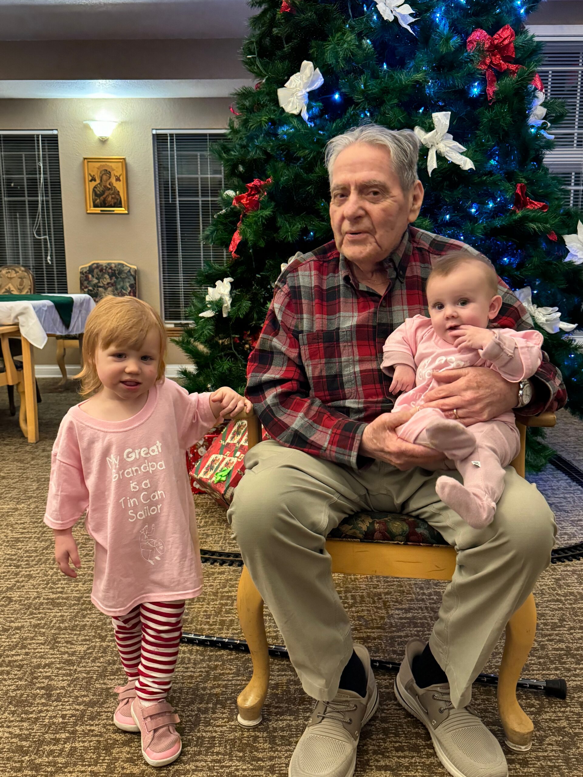 A grandfather sits in a chair holding a baby girl in pink, with a Christmas tree behind them and a loyal-looking toddler standing nearby in pink shirt.