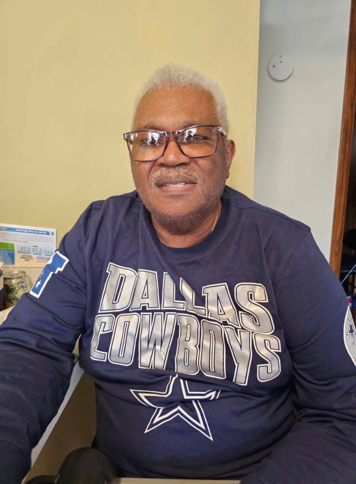Older man with gray hair and glasses wearing a Dallas Cowboys navy shirt, seated indoors against a light yellow wall.