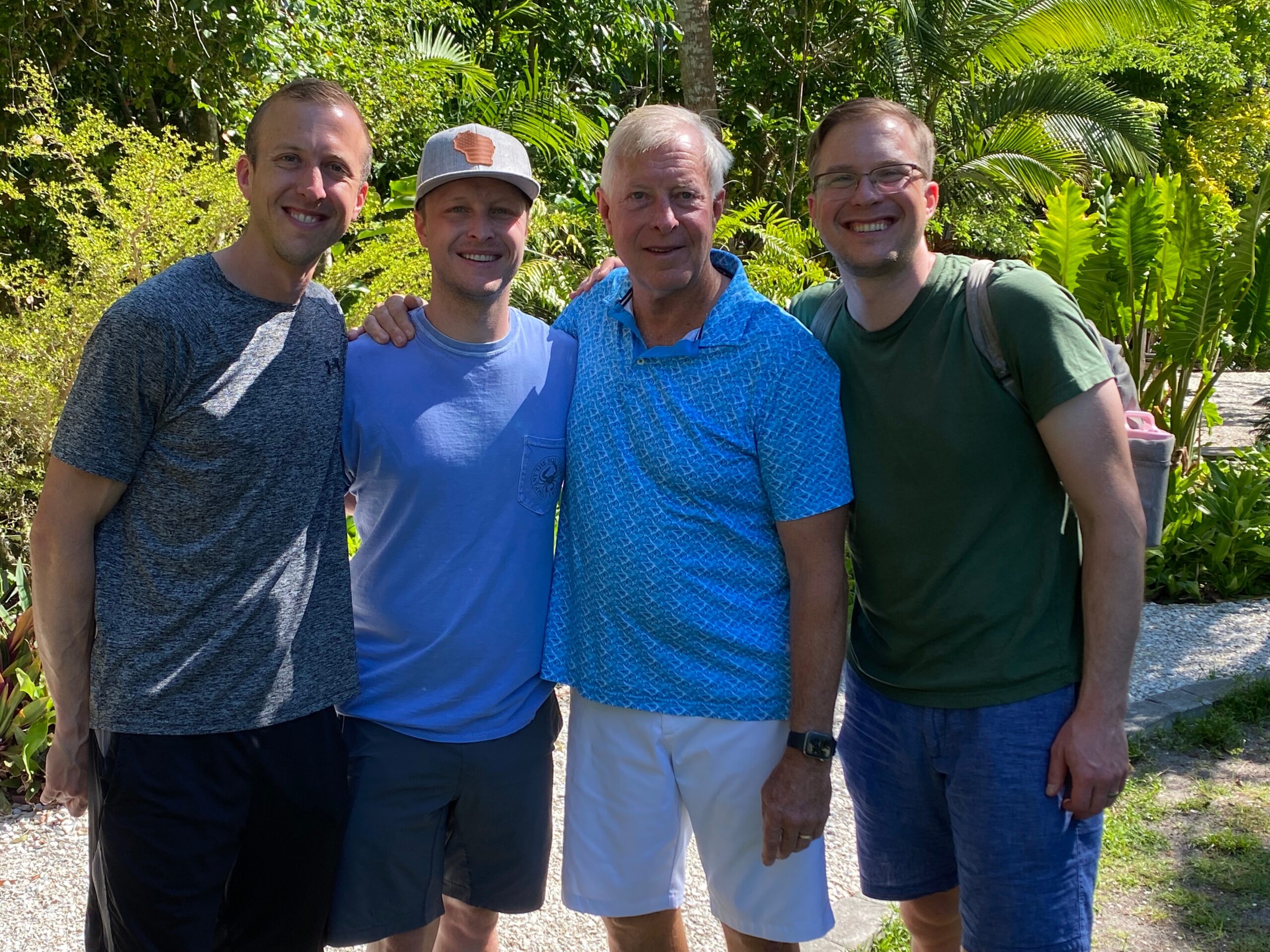 Four men stand together outdoors, smiling, with lush tropical plants in the background and arms around each other.