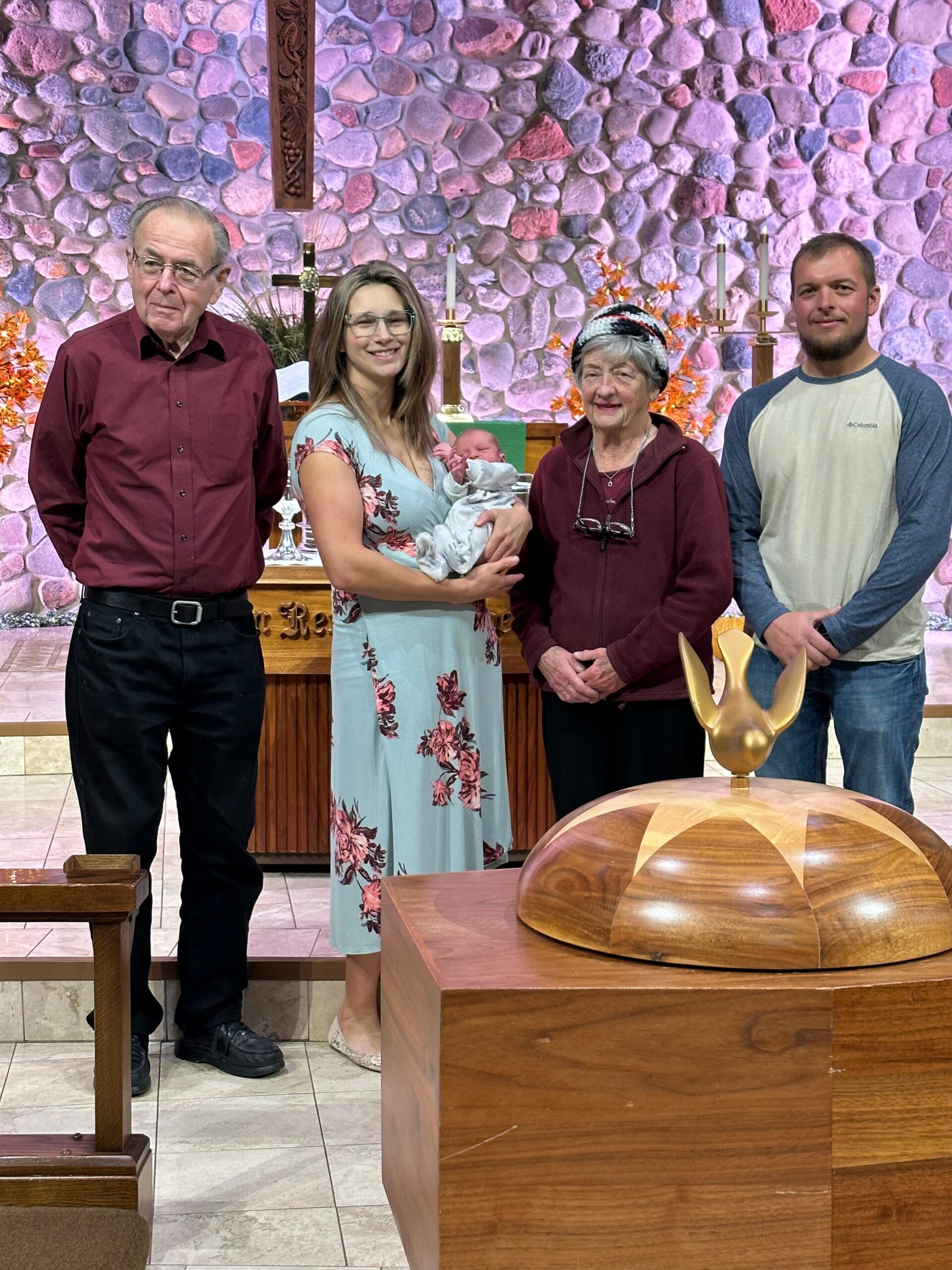 Family group posing at church altar, a woman in a light blue floral dress holds a newborn baby; two adults — an older man in a maroon shirt and an older woman in a burgundy jacket — stand with them, church stone wall backdrop behind with candles and cross visible.