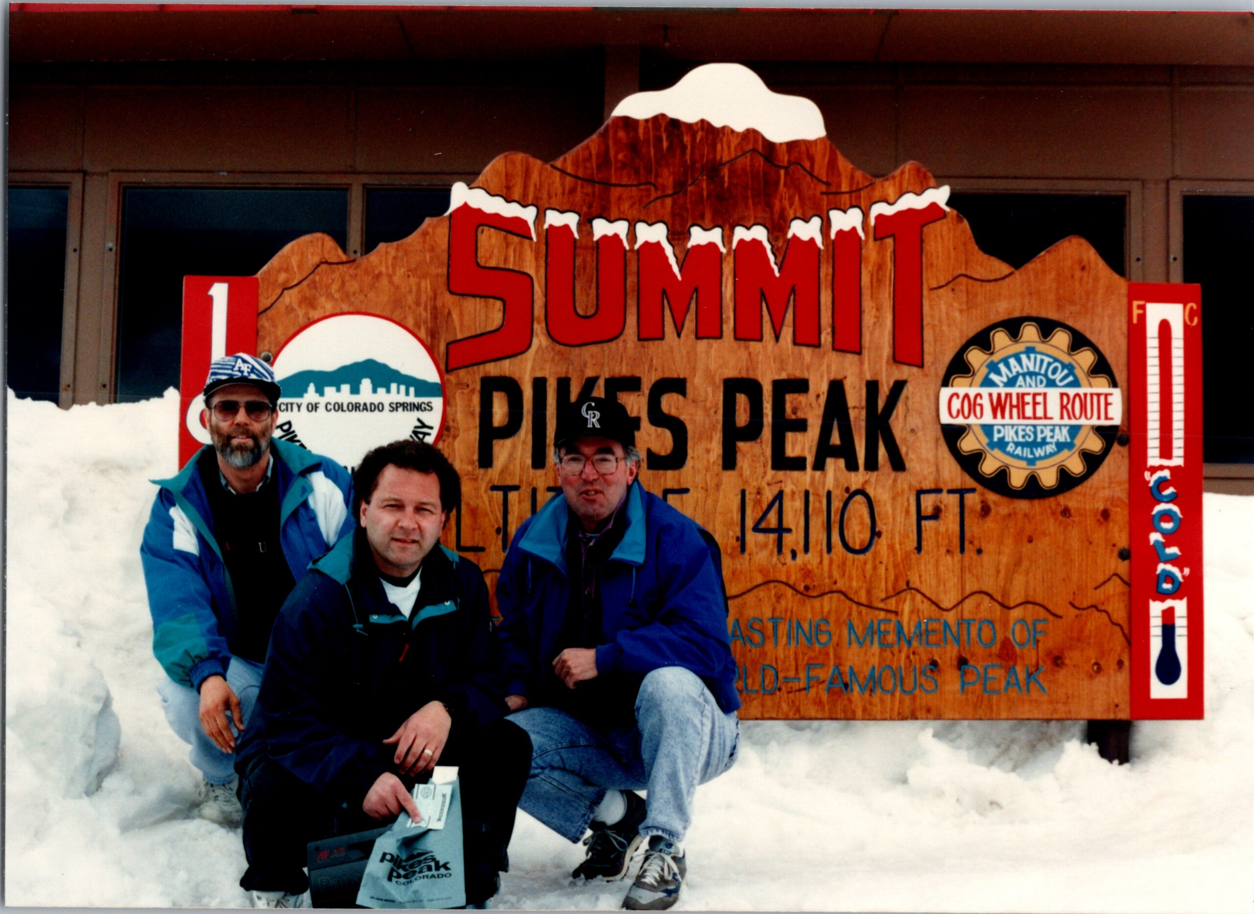 Group of four people in blue jackets posing in front of a large Summit Pikes Peak sign on a snowy day.