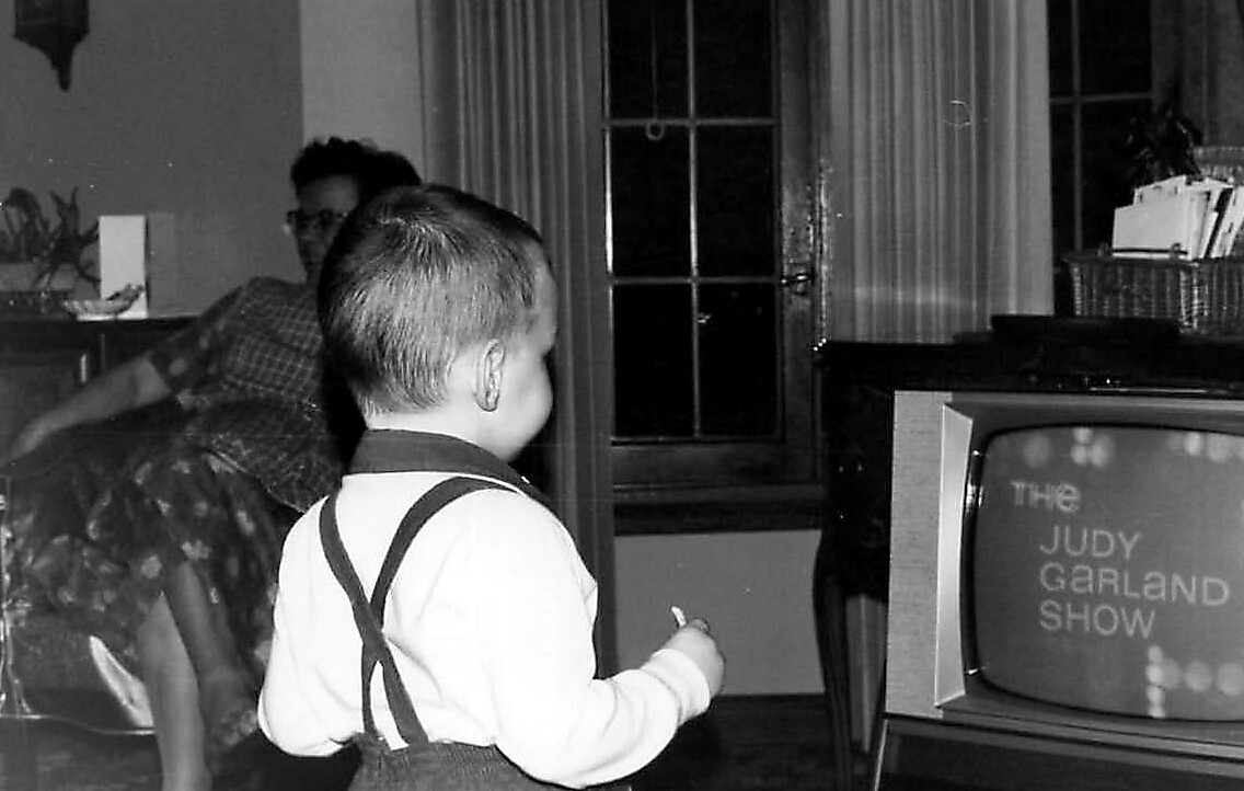 Young child with suspenders stands in a living room, facing a vintage TV that reads 'The Judy Garland Show'.