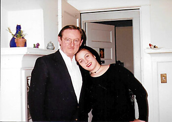 Couple posing together in a living room; woman leans her head on the man's shoulder, both smiling.