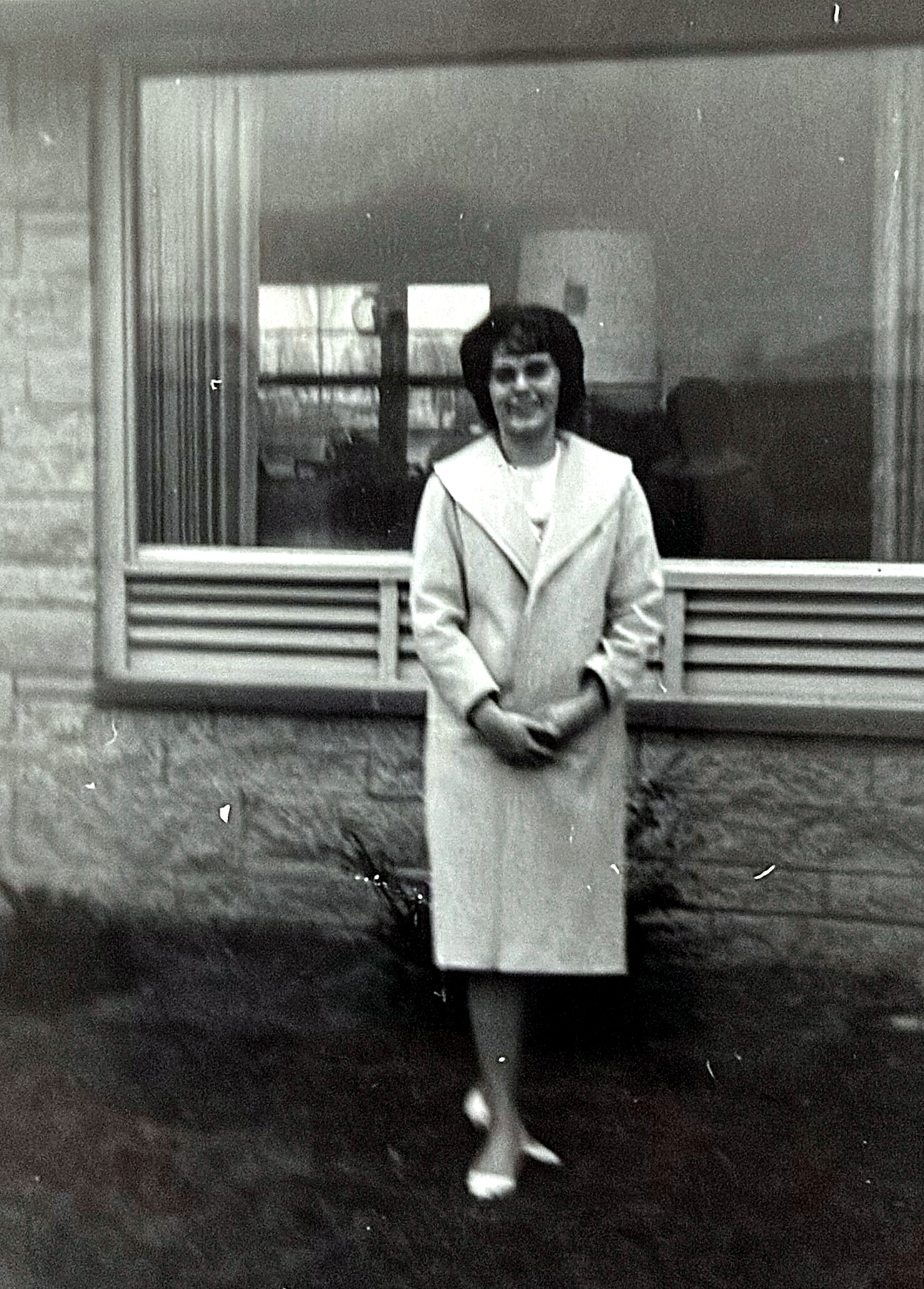 Smiling woman in a light coat standing outside in front of a large window, hands clasped.