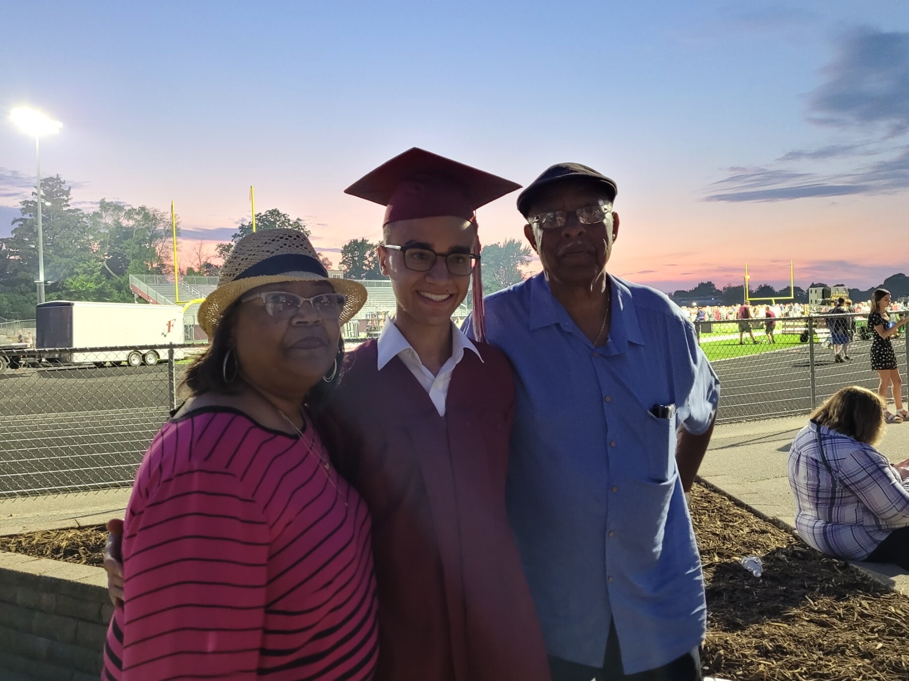 Three people pose at a football stadium at sunset: a woman in a pink striped top wearing a straw hat, a graduate in a maroon cap-and-gown, and a man in a blue shirt and cap.