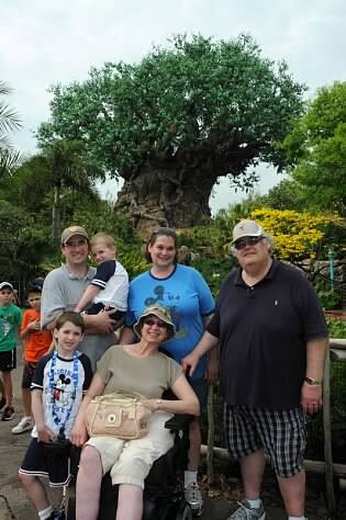 A group of adults and children posing for a photo in front of a large tree at a park; a woman in a wheelchair sits center with others standing around her.