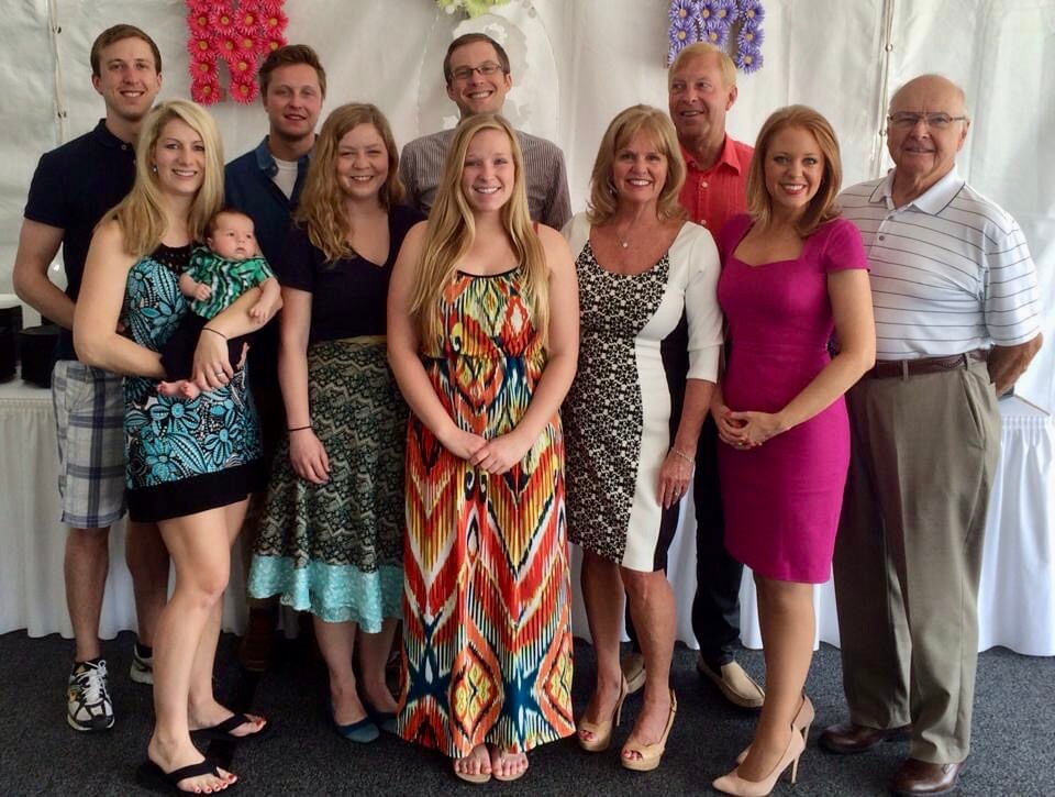 Group portrait of about 11 people (including a baby) posing together at a celebration, smiling in front of a floral backdrop.