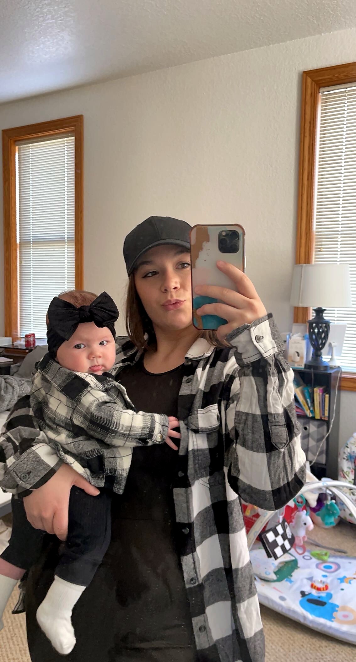 Woman in a black cap and black top, wearing a plaid shirt, holds a baby with a large black bow headband while taking a mirror selfie in a bright room.