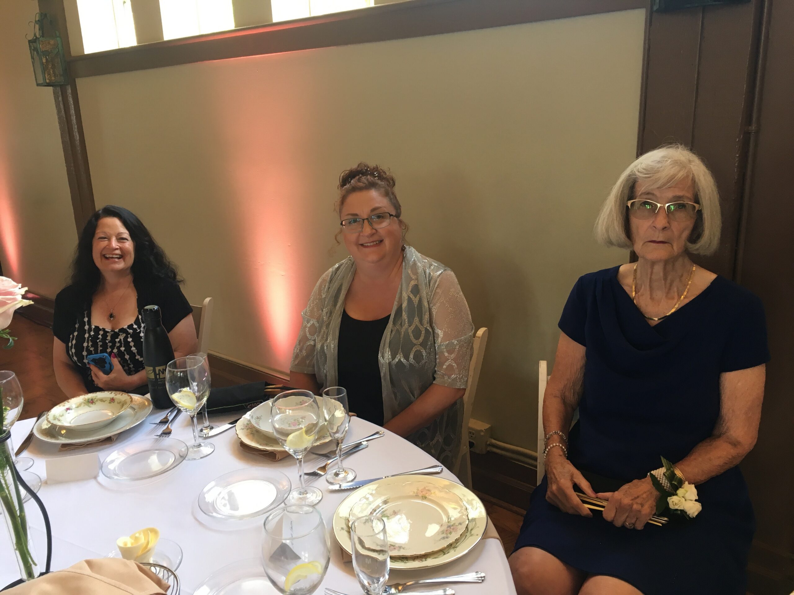 Three women seated at a dinner table with fine china and glasses, smiling during the meal.